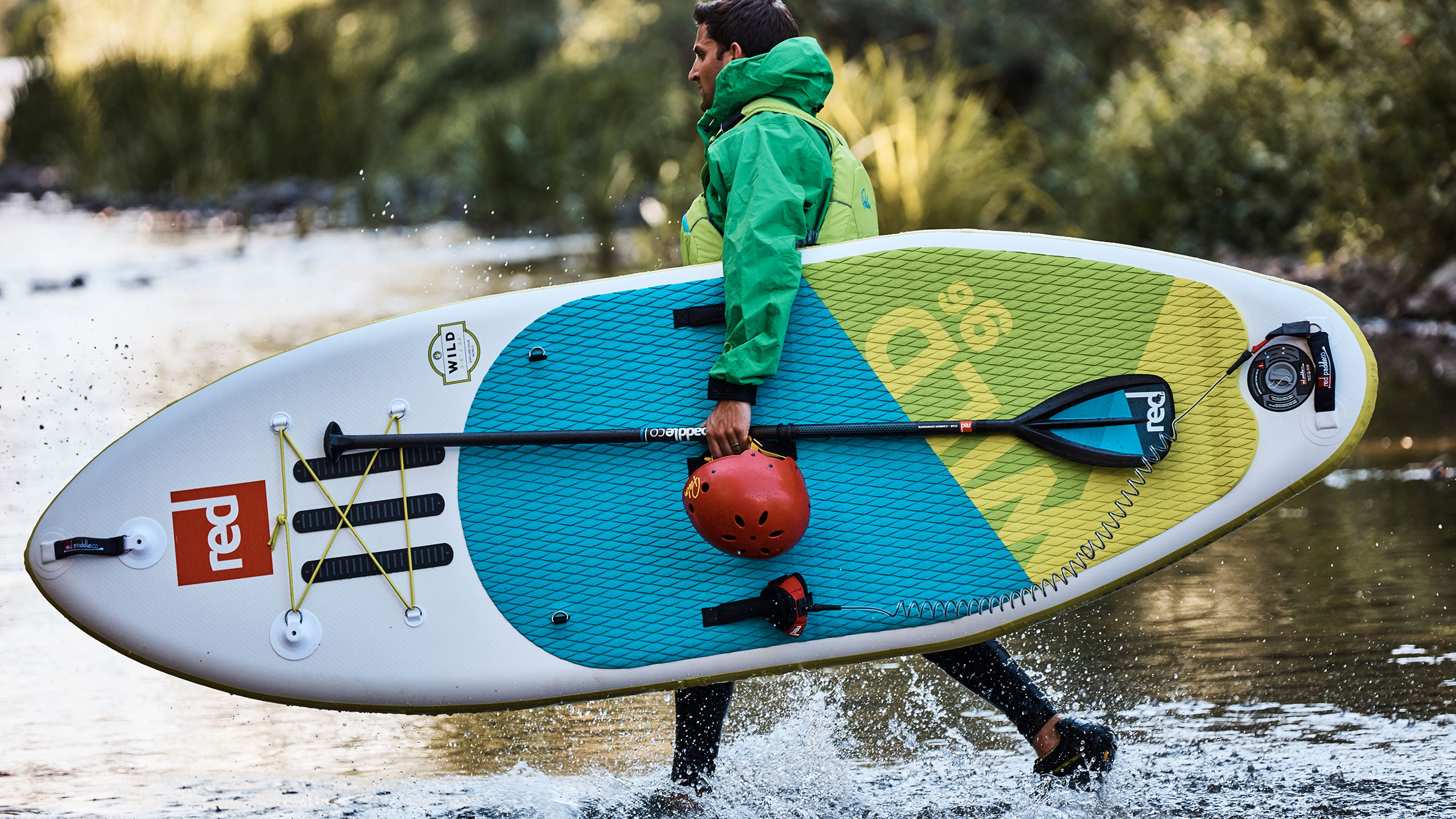 man walks through river carrying his Wild SUP, paddle and helmet