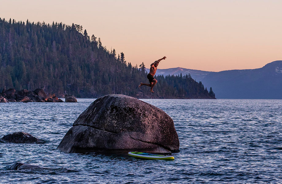 man jumps off rock with SUP on water below