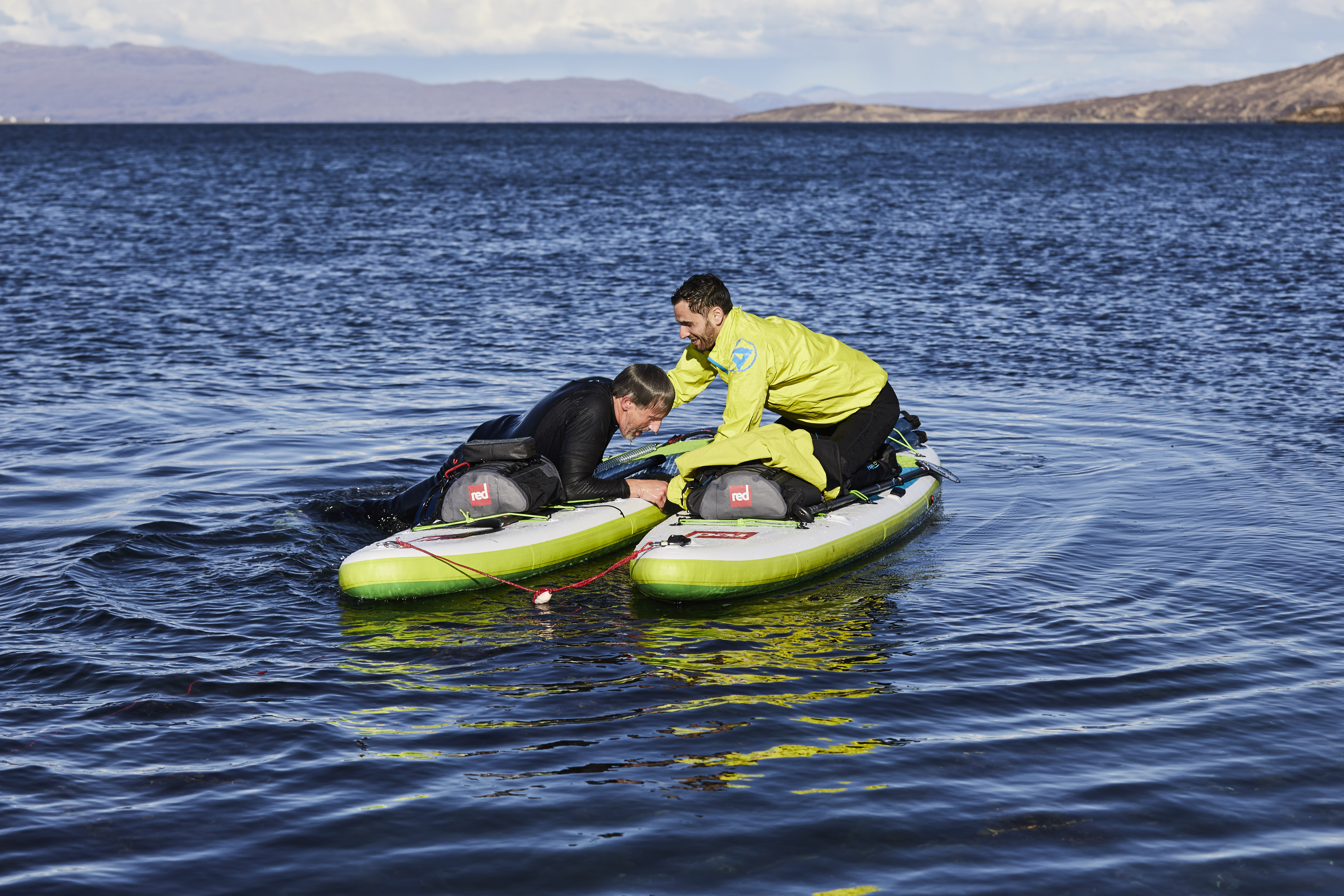 One male paddle boarder helps another back onto his inflatable paddle board after falling in